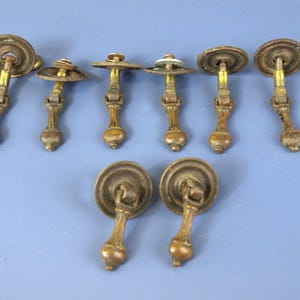 Op de afbeelding: Een verzameling antieke messing lade- en kastknoppen. De knoppen hebben een ronde achterplaat en een sierlijk, sierlijk ontwerp. De handgrepen hebben een vergelijkbaar ontwerp, met een afgeronde knop aan het uiteinde. Het beslag ligt op een lichtblauwe ondergrond.