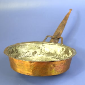 May include: A small, antique copper pan with a long, rusty handle. The pan has a rounded base and a slightly flared rim. The copper has a warm, reddish-brown hue, with areas of darker oxidation. The handle is attached with a simple bracket.