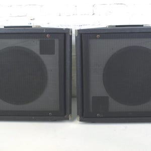 May include: Two black speaker cabinets with a mesh front and a large circular speaker cover. Each cabinet has a handle on top and a rectangular shape. The speakers are likely for a musical instrument or sound system.