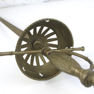Könnte beinhalten: Ein messingfarbenes Metallschwert mit einer langen, schlanken Klinge und einem detaillierten Griff. Der Griff ist mit einem breiten, kreisförmigen, perforierten Handschutz mit sonnenstrahlenartigem Muster und einer Parierstange verbunden.