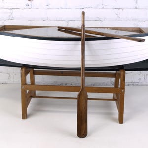 Puede incluir: Un bote de madera con casco blanco y ribete negro, apoyado sobre un soporte de madera. Un largo remo de madera está de pie frente al bote, con dos remos dentro. El bote mide aproximadamente 244 cm de largo.