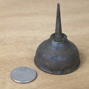 Può includere: Una lattina di olio vintage con un beccuccio lungo e appuntito. La lattina è in metallo e ha un colore marrone ruggine. È appoggiata su una superficie di legno.