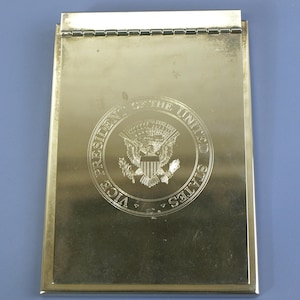 May include: A gold-colored metal clipboard with a hinged top. The words "VICE PRESIDENT OF THE UNITED STATES" and a detailed emblem are engraved on the front. The clipboard is rectangular with a smooth, reflective surface.
