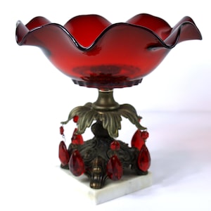 May include: A ruby red glass centerpiece bowl with a ruffled rim, supported by a bronze-toned metal base with leaf and teardrop crystal accents. The base rests on a white marble square.