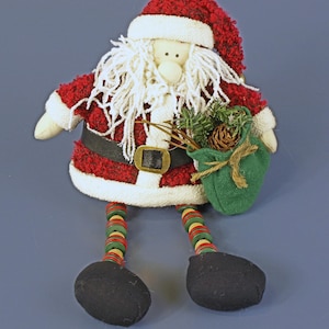 May include: A plush Santa Claus doll with a red and white suit, black belt, and black boots. The doll has a white beard and holds a green sack with pinecones and greenery. The legs are decorated with red, green, and yellow buttons.