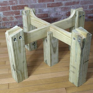 Puede incluir: Un soporte de madera para plantas con una parte superior en forma de cruz y cuatro patas robustas. El soporte está construido con madera de color claro y asegurado con tornillos. El diseño es simple y funcional, adecuado para exhibir plantas en macetas.