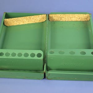 May include: Open, green metal tool case with two compartments. Each side has a row of holes and a cork strip. The word "GREENLEE" is visible on the front of each compartment. The case is open and empty.