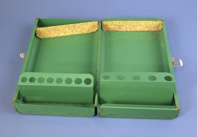 May include: Open, green metal tool case with two compartments. Each side has a row of holes and a cork strip. The word "GREENLEE" is visible on the front of each compartment. The case is open and empty.
