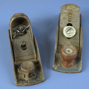 Puede incluir: Dos cepillos de mano de metal vintage, probablemente para carpintería. Los cepillos están desgastados, mostrando signos de edad y uso. Un cepillo tiene un pomo de madera. Los cepillos están sobre un fondo azul.
