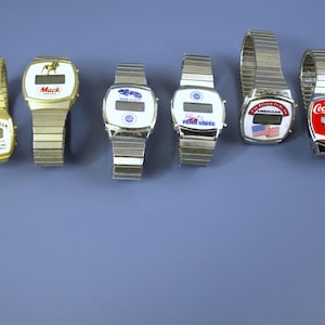 May include: A collection of eight silver and gold wristwatches with digital displays. The watches feature various brand logos, including Ford, Mack Trucks, Coca-Cola, and Pepsi.
