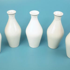 以下が含まれることがあります： 青い表面に配置された、光沢のある仕上げの白いセラミック花瓶が5つ。各花瓶は細い首、丸みを帯びたボディ、わずかにフレアした開口部を持っています。リムは濃い灰色のトリムでアクセントが付けられています。