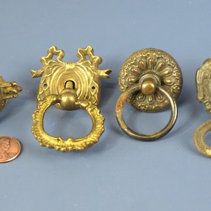 May include: Four antique brass drawer pulls with ornate designs and ring handles. The pulls are in various styles, including a floral design, a simple ring, and a more elaborate design with a ring handle.