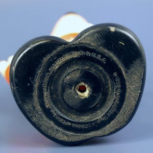 May include: A vintage toy figurine base, black and heart-shaped, with concentric circles and a central hole. The base has text that reads "Made in U.S.A." and "&copy; 1978 Gabriel CBS Toys, A Division". The figurine is white, orange, and black.