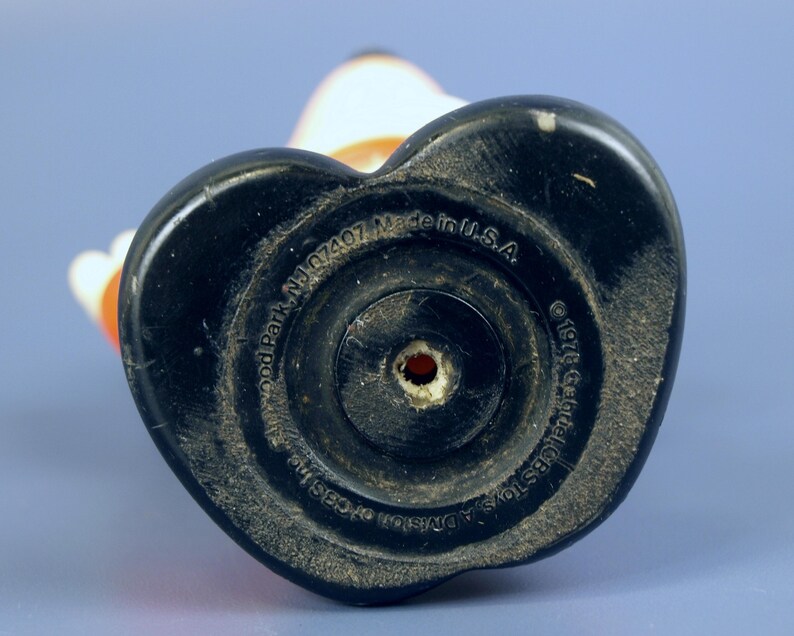 May include: A vintage toy figurine base, black and heart-shaped, with concentric circles and a central hole. The base has text that reads "Made in U.S.A." and "&copy; 1978 Gabriel CBS Toys, A Division". The figurine is white, orange, and black.