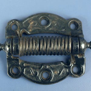 May include: An antique spring hinge, featuring a decorative design. The hinge is made of metal with a dark, aged finish. The spring is visible in the center, and the hinge has a decorative, ornate design. The hinge is designed to be mounted with screws.