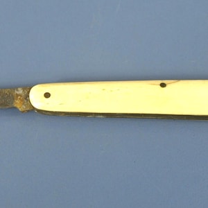 May include: A folding pocket knife with a weathered, metallic blade showing signs of oxidation and a smooth, cream-colored handle secured with two small metal rivets. The knife is shown in a closed position with the blade partially extended outward.