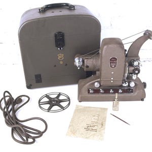 Puede incluir: Un proyector de películas Filmo marrón vintage con un estuche a juego, una bobina de película, un cable de alimentación y un manual de instrucciones. El proyector tiene varias perillas y botones. El estuche tiene un asa y un cierre metálico.
