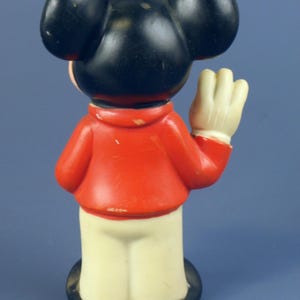 May include: A vintage Mickey Mouse figurine, viewed from the back. The figure has black ears and head, a red jacket, cream-colored pants, and white gloves. The base is black. The figurine is waving.