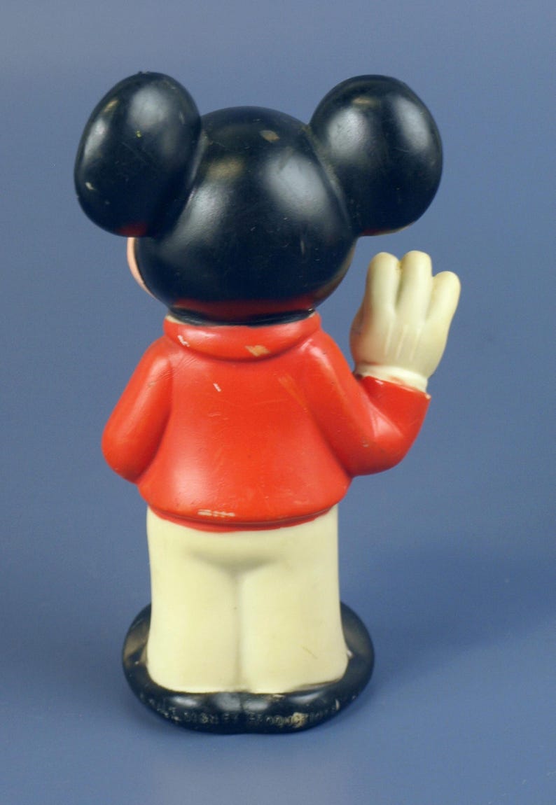 May include: A vintage Mickey Mouse figurine, viewed from the back. The figure has black ears and head, a red jacket, cream-colored pants, and white gloves. The base is black. The figurine is waving.