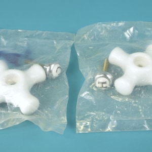 Peut inclure: Deux poignées de robinet blanches en forme de croix avec des accessoires métalliques sont emballées dans du plastique transparent. Les poignées ont une finition brillante et un trou central. Le fond est bleu clair uni.