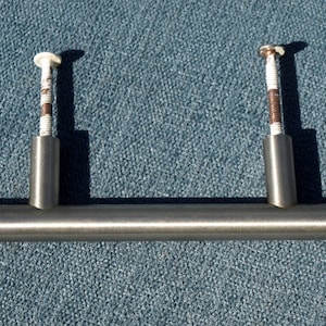 Puede incluir: Un tirador de armario de níquel cepillado con dos tornillos. El tirador es una barra recta, de aproximadamente 10 cm de largo, con postes cilíndricos para la fijación de los tornillos. Los tornillos tienen cabezas blancas y están roscados.