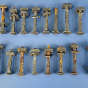Könnte beinhalten: Eine Sammlung antiker, verwitterter Metallbolzen und -befestigungen. Die Bolzen haben quadratische Köpfe und unterschiedliche Längen, mit sichtbarem Rost und Altersspuren. Einige enthalten Unterlegscheiben und Muttern, angeordnet auf blauem Untergrund.