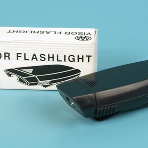 May include: A black visor flashlight and its white box. The box is labelled "VISOR FLASHLIGHT" and has a graphic of the flashlight. The flashlight is black and has a clip. The background is a light blue.