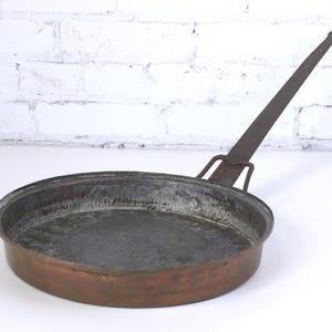May include: A vintage copper pan with a long, slender handle. The pan has a round, shallow cooking surface with a dark interior and a reddish-brown exterior. The handle is made of a darker metal and is attached to the pan with a bracket.