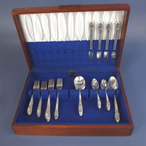 May include: A wooden silverware chest with a blue velvet interior and a white satin lining. The chest contains a set of silver-colored forks, spoons, and knives. The silverware is arranged in individual slots within the chest.
