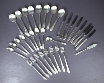 May include: A collection of vintage silverware, including spoons, forks, knives, and a butter knife, arranged on a dark gray surface. The silverware has a floral design on the handles and a shiny, metallic finish. The arrangement suggests a formal dining setting.