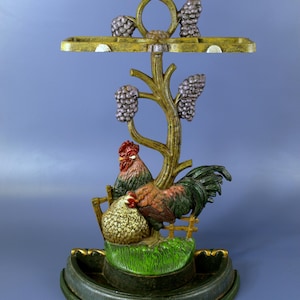 May include: Cast iron umbrella stand with a rooster and hen design. The stand features a green base, a green and gold tree with purple grapes, and a gold top with a tray for umbrellas.