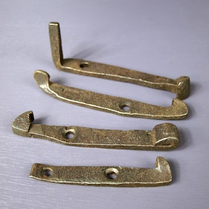 May include: Four antique bronze-colored metal latches. The latches have a rustic, aged appearance with visible wear and tear. Each latch features a curved or angled design with pre-drilled holes for mounting. The metal has a slightly textured surface.