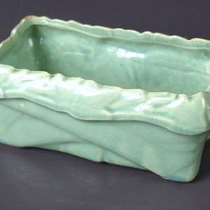 以下が含まれることがあります： 長方形で、テクスチャのある折り畳まれたデザインの薄い緑色のセラミックプランター。プランターは、わずかに波打った縁を持っています。