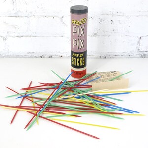 Può includere: Un gioco vintage di Pick-Up Sticks con un contenitore cilindrico rosa e nero. Il contenitore recita "Plastic Pix Pix Pick-Up Sticks". Un mucchio di bastoncini colorati in rosso, blu, verde e giallo sono sparsi.