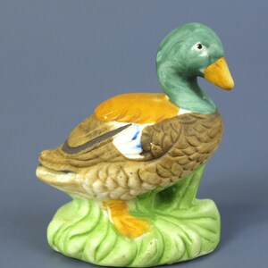 May include: A ceramic duck figurine with a green head, orange chest, and brown wings. The duck has a yellow beak and is perched on a green, leaf-shaped base. The figurine is painted with detailed feather textures.