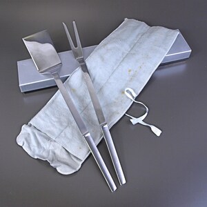 May include: A stainless steel barbecue set including a spatula and a carving fork, resting on a light blue cloth. The set is presented with a silver box and a white ribbon. The utensils have long, sleek handles.