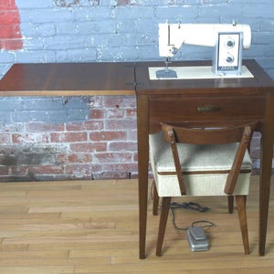 May include: A vintage sewing machine on a wooden cabinet with a pull-out extension. The machine is white and silver, with a foot pedal on the floor. A small chair with a cushioned seat is positioned in front of the cabinet.