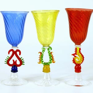 May include: Three decorative wine glasses with coloured bowls and clear bases. The bowls have a spiral design. The stems feature ornate, colourful glass designs in red, yellow, and blue. Each glass has a clear, flared base.