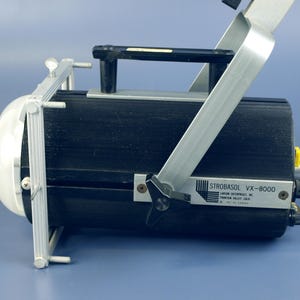 May include: A black and silver strobe light, model VX-8000, with a white dome and a handle. The light has a silver frame and is on a blue surface. The text "STROBASOL VX-8000" is visible.
