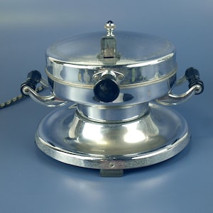 Puede incluir: Una gofrera cromada vintage con asas negras y una base decorativa. El aparato tiene una tapa con bisagras con un pequeño asa y un dial. La gofrera está diseñada para hacer gofres.