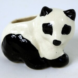 Może przedstawiać: Ceramiczna figurka pandy, pomalowana na czarno-biało, z małym otworem na plecach. Panda ma czarne uszy, oczy i nogi oraz białe ciało. Ten element dekoracyjny to uroczy dodatek do każdej kolekcji.