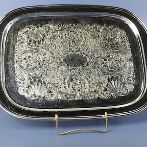 May include: A silver-coloured, rectangular serving tray with ornate, raised floral and scrollwork detailing. The tray has a dark border and is supported by a gold-coloured metal stand. The tray's surface reflects light, highlighting its intricate design.