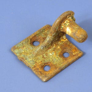 Puede incluir: Un gancho y una placa de metal oxidados y desgastados, con tres agujeros. El gancho se extiende desde una placa cuadrada, mostrando signos de edad y oxidación. El metal tiene una mezcla de tonos marrones, verdes y dorados.
