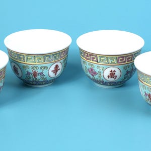 Könnte beinhalten: Vier weiße Porzellan-Teetassen mit einem türkisfarbenen, gelben und roten Zierband. Jede Tasse hat ein Blumendesign und chinesische Schriftzeichen. Die Tassen sind auf einer hellblauen Oberfläche angeordnet.