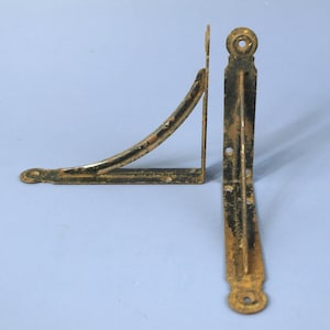 May include: Two L-shaped metal shelf brackets with a distressed black and gold finish. Each bracket features a curved support brace, flat mounting arms, and circular screw holes at the ends and along the base for secure wall attachment.
