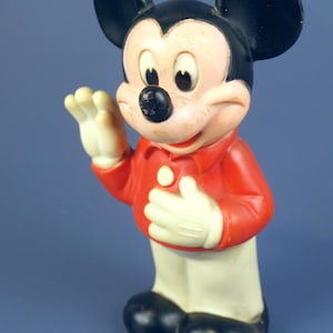 May include: Vintage Mickey Mouse figurine. The figure is wearing a red shirt, white gloves, and white pants. The iconic character has black ears, a black nose, and a smiling expression. The toy is standing and waving.