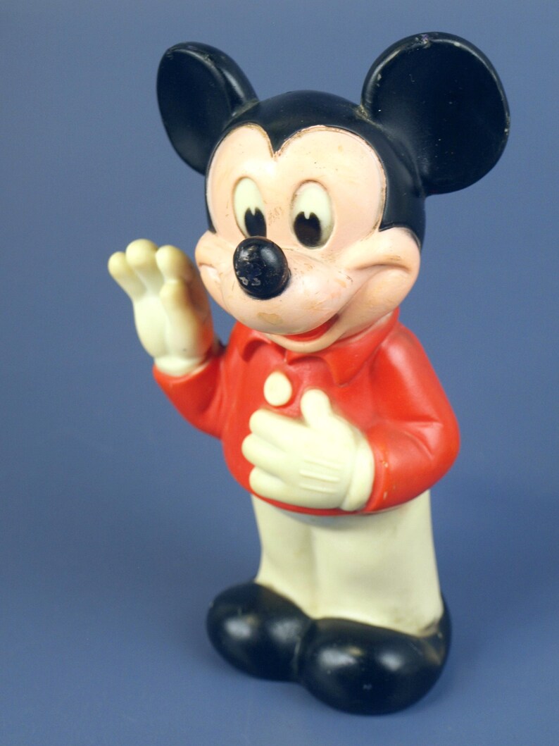 May include: Vintage Mickey Mouse figurine. The figure is wearing a red shirt, white gloves, and white pants. The iconic character has black ears, a black nose, and a smiling expression. The toy is standing and waving.