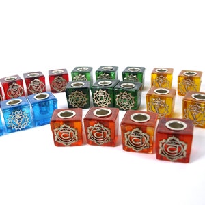 May include: Set of 18 colorful glass candle holders with silver chakra symbols. Each holder is a cube with a small opening at the top for a tea light candle.