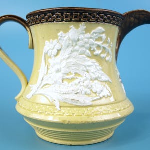 May include: A ceramic pitcher featuring a pale yellow body with raised white floral relief patterns. The rim and handle are finished in a dark brown glaze, and the upper rim displays a decorative geometric border pattern.