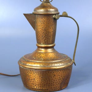 May include: A hammered copper lamp base shaped like a traditional pitcher or ewer. The metal surface features a distinct dimpled texture throughout the body, a curved handle on the side, and a pointed spout. A lamp socket is attached to the top.